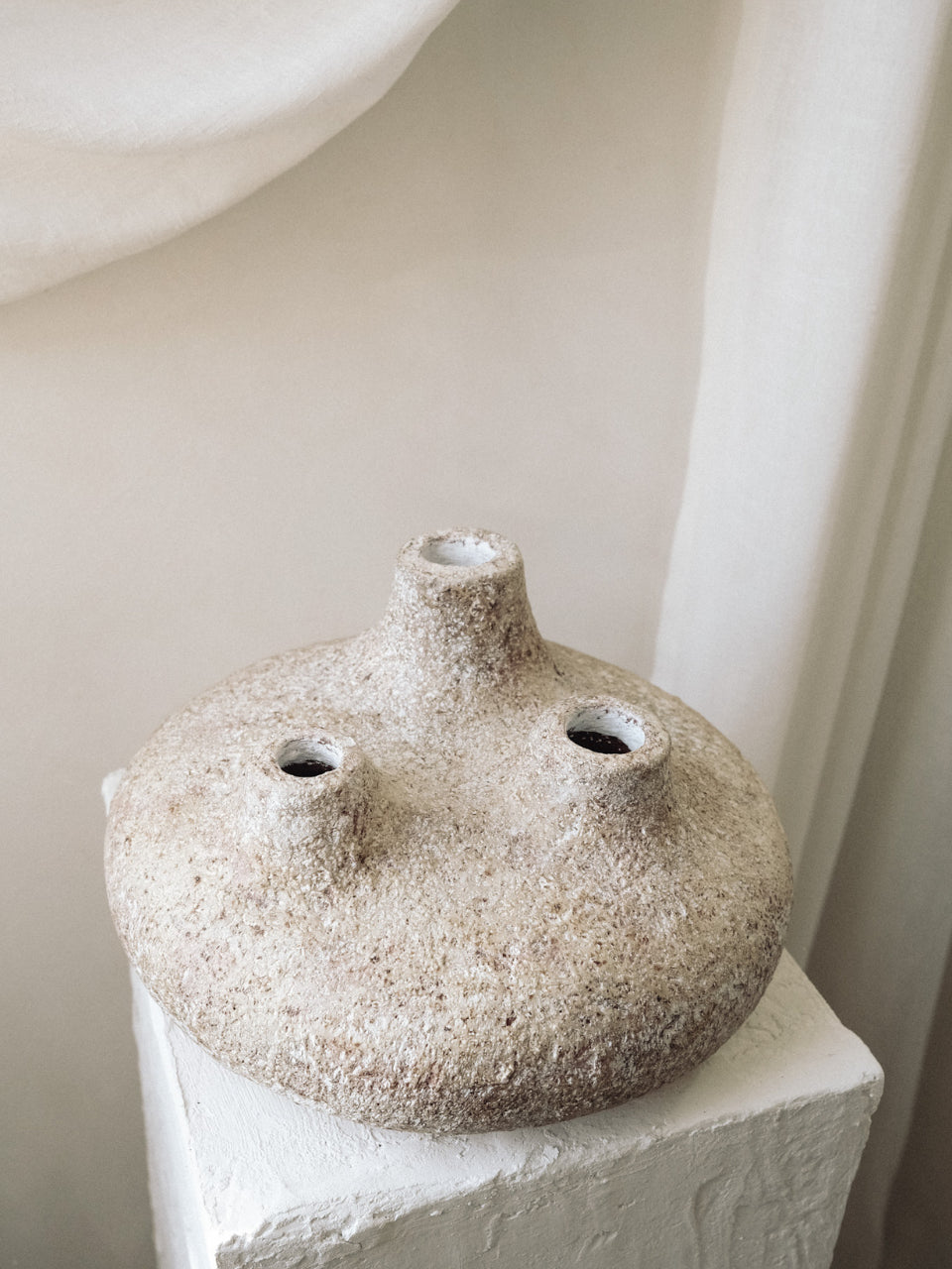 Amit three-spout terracotta vessel on a white pedestal against soft draped linen