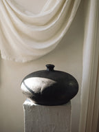 Dark Rovi terracotta moon vessel with a low rounded body on a white plaster pedestal