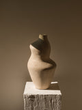 Handmade Suma terracotta vessel with a curved sculptural silhouette in matte cream on a white plaster pedestal