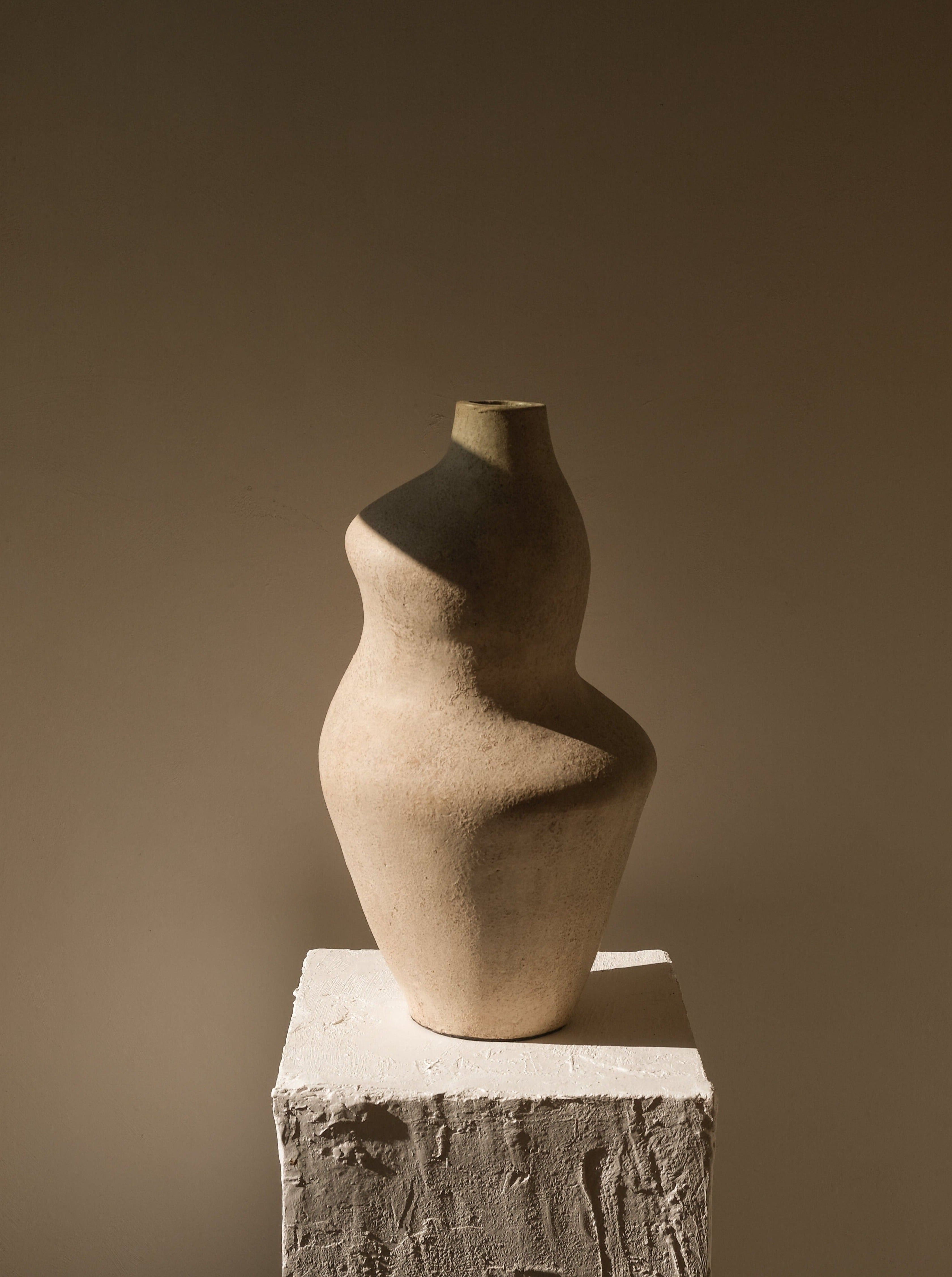 Handmade Suma terracotta vessel with a curved sculptural silhouette in matte cream on a white plaster pedestal