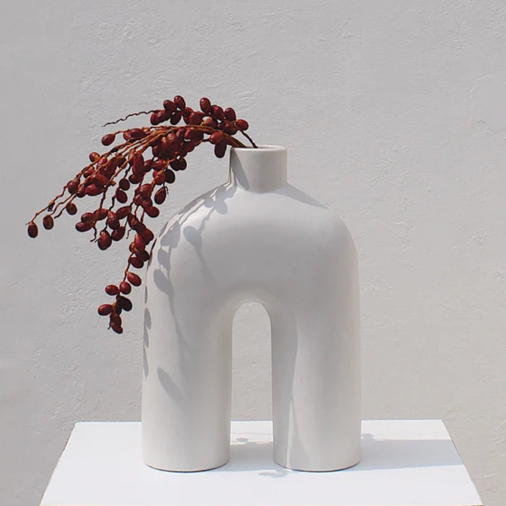 Off white Ozo ceramic arch vase holding a draping red berry branch on a white plinth