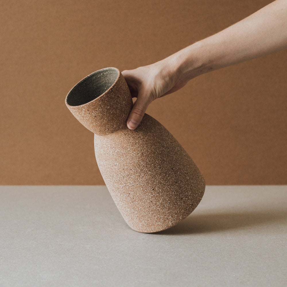 Hand tilting the Reloj ceramic vase to show its cinched hourglass form and raw clay texture