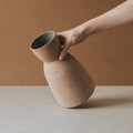 Hand tilting the Reloj ceramic vase to show its cinched hourglass form and raw clay texture