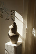 Suma vessel holding a tall budding branch on a plaster pedestal against draped linen