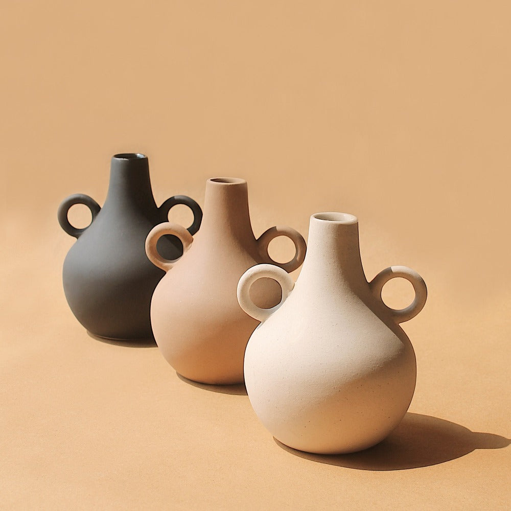 Three Harappan ceramic jug vases in charcoal, tan, and cream grouped on a warm tan backdrop