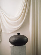  Dark sculptural moon vase with small flat-top neck against a draped curtain
